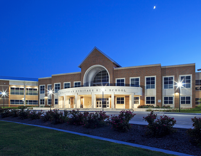 A brick high school at dusk near Dallas, with lights on and a crescent moon in the sky.